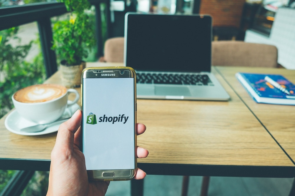 Photo is of a hand holding a smartphone displaying the Shopify logo in front of a café table with a coffee.