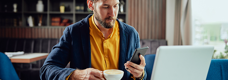 Young Caucasian bearded businessman dressed smart casual using a smart phone and holding a cup of coffee.