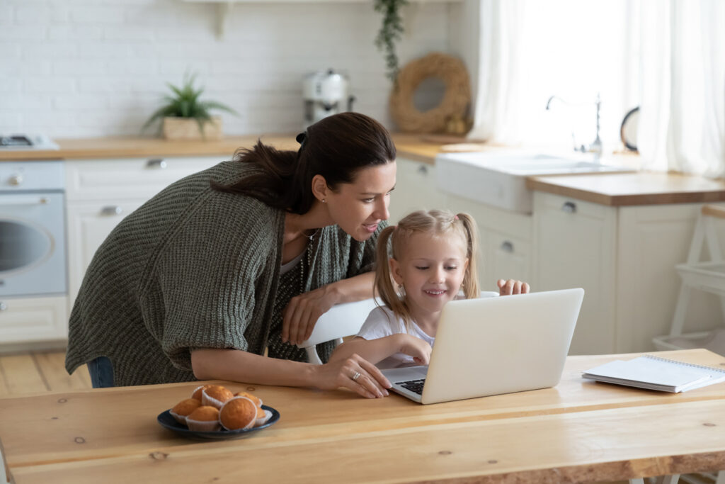 A mother and daughter are looking at a laptop on a wooden table, in a bright, modern kitchen. A plate of rolls is nearby. The kitchen has white walls and natural light coming from the window. An open notebook is also visible beside the laptop.
