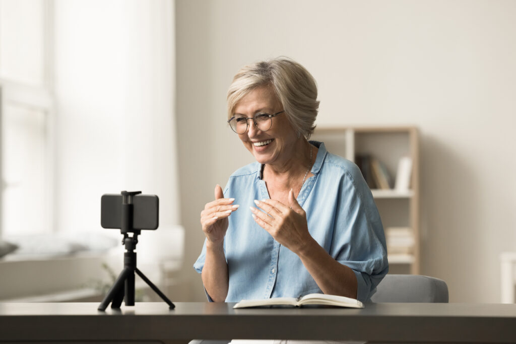 An older woman sitting at a table, smiling at a device, recording a video or live stream, using a smartphone mounted on a tripod, while gesturing and speaking into the camera in an indoor setting.
