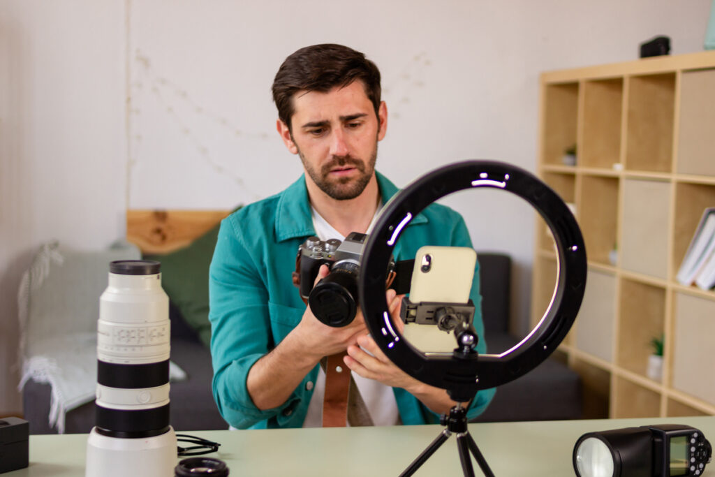 A person with their face obscured is holding a camera, with a ring light illuminating a smartphone mounted on a tripod; various photography equipment is scattered around.